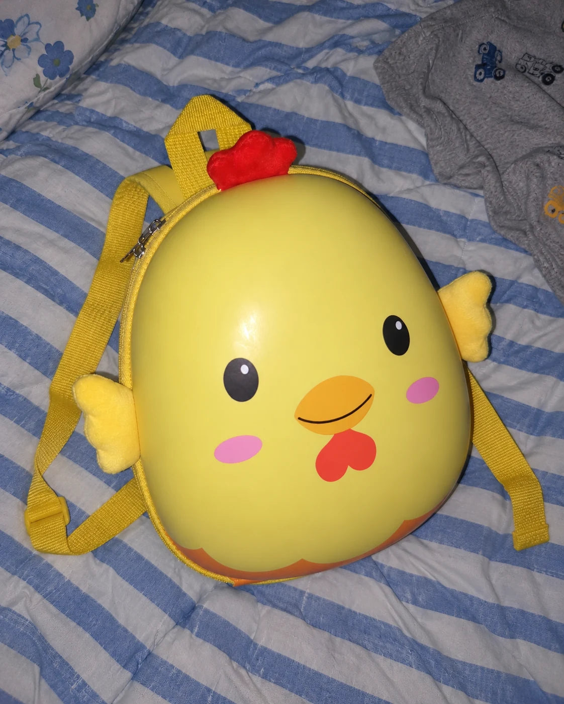 Illustrative overhead phone-style photo of the yellow chick backpack on a bed